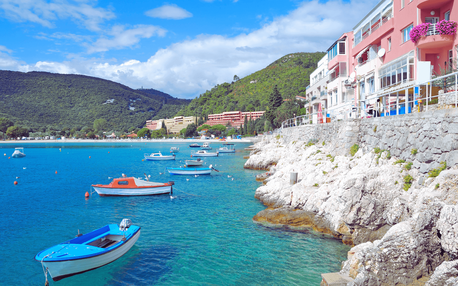 A serene coastal scene of Medulin beaches for all ages, featuring clear blue waters with boats, rocky shores, and colorful buildings along the coastline. Green hills and a partly cloudy sky enhance the inviting atmosphere for families and seniors alike.