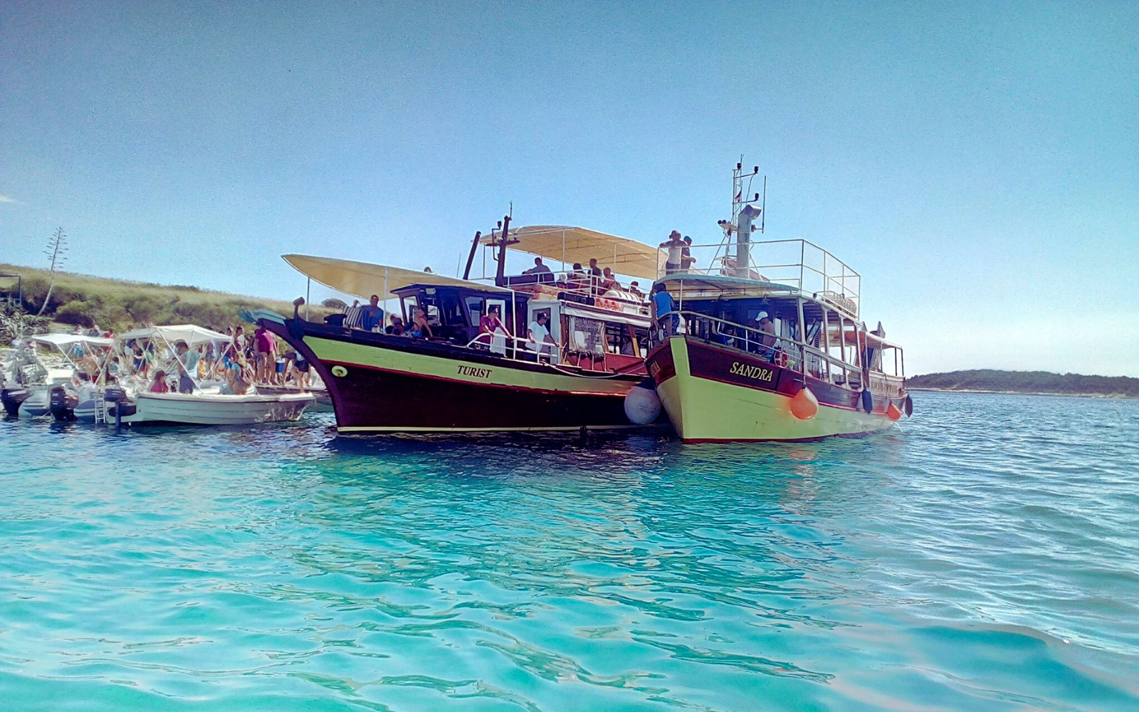 Two boats, "Turist" and "Sandra," filled with people on clear turquoise waters, showcasing some of the best Medulin activities for families. The scene includes a sunny day with green hills, offering a fun and scenic boat trip experience for all ages.